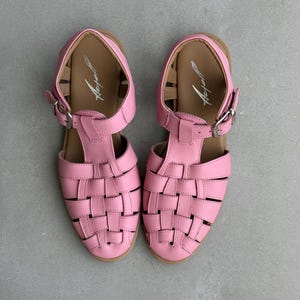 K&ouml;nnte beinhalten: Ein Paar rosa Ledersandalen mit einem gewebten Design auf der Vorderseite. Die Sandalen haben einen Schnallenverschluss und eine braune Innensohle. Die Sandalen sind etwa 23 cm lang.