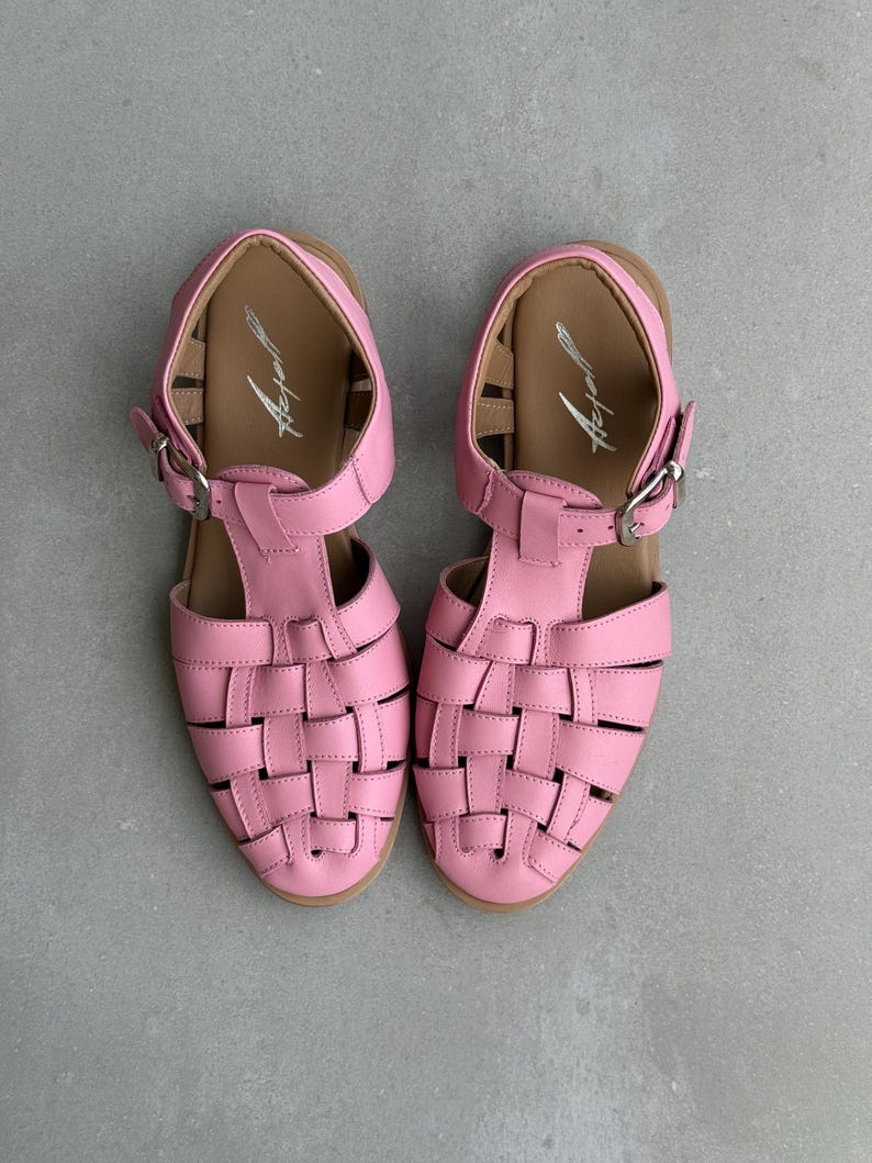 K&ouml;nnte beinhalten: Ein Paar rosa Ledersandalen mit einem gewebten Design auf der Vorderseite. Die Sandalen haben einen Schnallenverschluss und eine braune Innensohle. Die Sandalen sind etwa 23 cm lang.