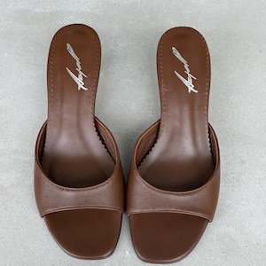 May include: A pair of brown open-toe mules with a low heel. The footbed features a smooth brown surface with a silver script logo printed near the heel area. The design includes a wide front strap and a rounded toe shape, showcasing a clean finish.