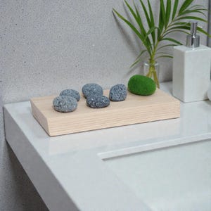 May include: A pale wooden tray displays grey stones and a green moss ball. A white soap dispenser and a small glass vase with green leaves are in the background. The items are arranged on a white countertop.