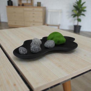 May include: A black, organically shaped tray holds several grey stones and a green, moss-like object. The tray sits on a light-coloured wooden table, creating a minimalist, zen-like aesthetic. The tray's design and contents suggest a focus on natural elements.