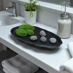 May include: A black, oval decorative tray with green moss and grey stones. The tray is on a white countertop next to a small potted plant and a glass jar with a metal top. A sink and towels are also visible.