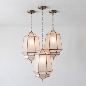 Pode incluir: Um candeeiro de teto com três luzes, apresentando abajures geométricos em forma de lanterna com painéis de vidro fosco e uma estrutura de metal em tom de bronze. Cada luz pende de uma corrente a condizer e de uma base circular.