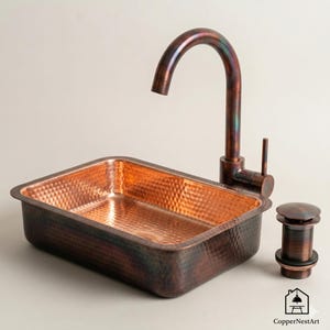 Könnte beinhalten: Ein kupfernes Waschbecken mit gehämmerten Texturen, eine passende Armatur und ein Ablaufstopfen. Das Waschbecken ist rechteckig und hat eine satte, rotbraune Farbe. Die Armatur hat einen gebogenen Auslauf. Der Ablaufstopfen ist zylindrisch.