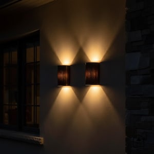 May include: Two outdoor wall sconces with a dark brown textured shade, mounted on a light-colored wall. The sconces emit a warm, downward-facing light, creating a dramatic effect. The scene is set at night, with a dark background.