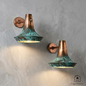 May include: Two copper wall sconces with a verdigris finish. The sconces have a bell-shaped shade and a circular wall mount. The sconces are mounted on a gray wall. The sconces are lit.