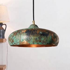 May include: A dome-shaped pendant light fixture featuring a hammered metal exterior with a weathered patina of oxidized green and copper tones. The interior of the shade has a warm, reflective metallic finish, suspended by a black cord.