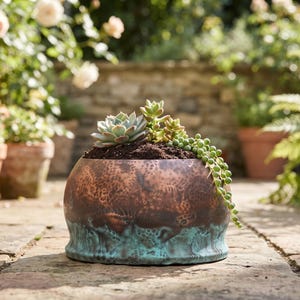 Pode incluir: Um vaso redondo de cobre martelado com um acabamento de pátina turquesa na base. O vaso está cheio de terra escura e uma variedade de suculentas, incluindo uma echeveria em forma de roseta e uma planta de colar de pérolas pendente.