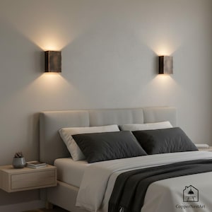 May include: Two bronze-colored wall sconces illuminate a bedroom. Each rectangular sconce emits a warm glow, casting light upwards and downwards. The sconces are mounted above a bed with gray pillows and a light-colored headboard.