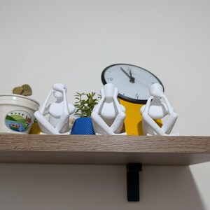 May include: Three white minimalist sculptures of figures in the "See No Evil, Hear No Evil, Speak No Evil" pose. They sit on a wooden shelf with a clock and potted plants in the background. The shelf is supported by a black metal bracket.