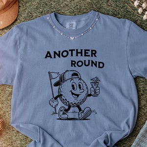 May include: A blue t-shirt with the text "ANOTHER ROUND" above a cartoon golf ball character holding a cocktail and a golf flag. The shirt has a crew neck with a decorative necklace. The bottom right corner of the shirt has the text "BlueJean".