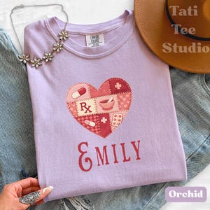 Puede incluir: Una camiseta de color lila claro con un diseño en forma de corazón con símbolos médicos y el nombre "EMILY" en rojo. La camiseta está hecha de material Comfort Colors. La palabra "Orchid" está impresa en la parte inferior derecha.