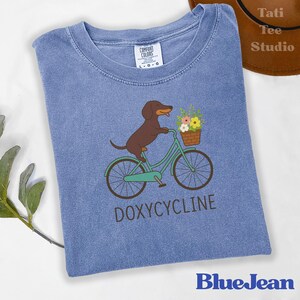 以下が含まれることがあります： 水色のTシャツ。茶色のダックスフンドが、花のバスケットを載せたターコイズ色の自転車に乗っているイラスト。下に「DOXYCYCLINE」の文字。ブランド名「BlueJean」が下部に印刷されています。