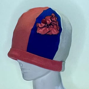 May include: A color-blocked beanie hat with sections of coral, blue, and white. The hat features a coral ribbed brim and a decorative red fabric flower on the blue panel. The hat is displayed on a white mannequin head.
