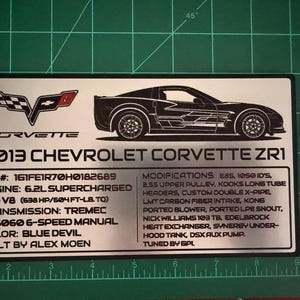 May include: A silver metal plaque featuring a black outline of a Corvette ZR1 car. The plaque includes the Corvette logo, the text "2013 Chevrolet Corvette ZR1", and detailed specifications such as engine, transmission, and modifications.