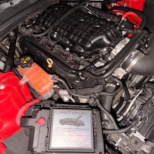 May include: A detailed view of a car engine compartment. The engine is black with various tubes and wires. A red coolant reservoir and red wiring are visible. A black box with the word "CAMARO" and text is in the foreground.