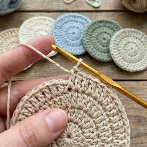 May include: Close-up of crochet work in progress. A golden crochet hook is inserted into a beige crocheted circle. Several other crocheted circles in various pastel colors are in the background. The hook has the word "DCeret" printed on it.