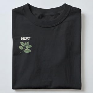 May include: Black t-shirt with a crew neck. The shirt displays the word "MINT" in white, above a green illustration of mint leaves and stems. The t-shirt is neatly folded.
