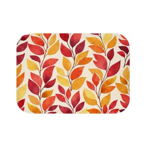 May include: A rectangular bath mat featuring a repeating watercolor pattern of autumn leaves in shades of deep red, bright orange, and golden yellow against a cream-colored background. The leaves are arranged on thin, dark brown stems.