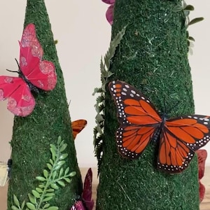 Puede incluir: Dos decoraciones en forma de cono cubiertas de musgo verde, adornadas con mariposas coloridas y vegetación. Las mariposas son en tonos de rosa, naranja y morado. Los conos están sobre un fondo neutro.