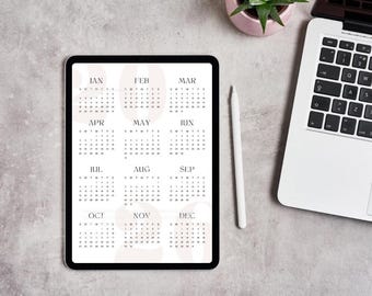 May include: A digital calendar displayed on a tablet, showing the months of the year. The tablet is on a light gray surface, next to a white stylus, a laptop, and a small potted plant. The calendar has a minimalist design.