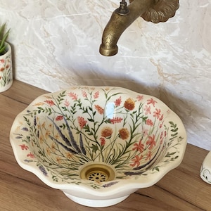 Könnte beinhalten: Ein mit Blumen verziertes Keramikwaschbecken mit einer Messingarmatur. Das Waschbecken hat einen gewellten Rand und ist mit einem farbenfrohen Wildblumenmuster verziert. Ein kleiner Blumentopf und eine Seifenschale mit passenden Blumenmustern sind ebenfalls zu sehen.