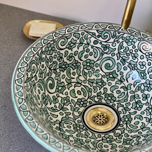 Peut inclure: Un lavabo en céramique décoratif avec un motif floral vert et blanc. Le lavabo est de forme ronde avec un bord décoratif et un drain doré. Un porte-savon doré avec un savon est visible en arrière-plan. Le diamètre est d'environ 30 cm.