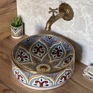 Puede incluir: Un lavabo de cerámica decorativo con un colorido estampado floral en azul, amarillo, rojo y verde. El lavabo tiene un borde y un desagüe de color dorado. Un grifo de latón de estilo vintage está montado en la pared sobre el lavabo. A la izquierda, una pequeña maceta de cerámica con una planta.