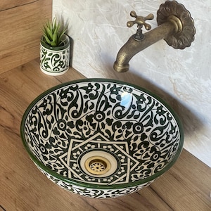 May include: A decorative ceramic sink with a green and white floral pattern. The sink has a round shape and a brass drain. A brass faucet and a small decorative plant in a matching pot are also visible.