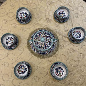 Könnte beinhalten: Ein Keramik-Teeservice mit einem dekorativen Blumenmuster in Rot, Blau und Gelb. Das Set enthält sechs Tassen und Untertassen sowie eine Servierschale mit Deckel. Die Ränder der Tassen und Untertassen sind helltürkis.