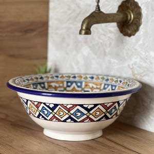 Può includere: Un lavabo decorativo in ceramica con base bianca e bordo blu. Il lavabo presenta un colorato motivo geometrico in rosso, blu, giallo e verde. Un rubinetto vintage in ottone è montato sulla parete dietro il lavabo.