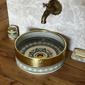 Può includere: Un lavabo decorativo in ceramica con bordo dorato e intricati motivi verdi e bianchi. Il lavabo è rotondo e poggia su una superficie in legno, con un portasapone e un portaspazzolino abbinati. Un rubinetto in stile vintage è montato a parete.
