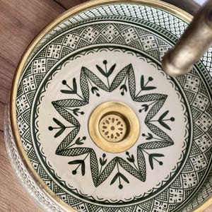 Könnte beinhalten: Ein dekoratives Keramikwaschbecken mit cremefarbenem Innenraum und grünem geometrischen Muster. Das Becken hat einen goldfarbenen Rand und einen goldfarbenen Abfluss. Das Design zeigt ein zentrales sternförmiges Motiv und ein sich wiederholendes Bordürenmuster.