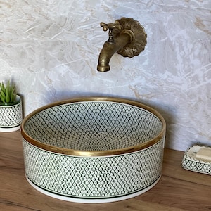 Peut inclure: Un lavabo de salle de bain décoratif avec un motif géométrique vert et blanc, accentué d'un bord doré. Un robinet en laiton vintage est monté sur le mur au-dessus. Un porte-savon assorti et un petit pot de plante sont également visibles.