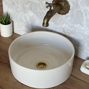 Peut inclure: Un évier rond en céramique blanche avec un drain et un robinet en laiton. Le robinet a un design orné. Une petite plante en pot et un porte-savon sont visibles. L'évier est posé contre un mur à motifs de marbre.