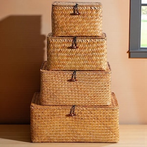 May include: Four woven rectangular storage baskets of varying sizes are stacked. The baskets are made of a light brown material and have lids with a dark string and wooden toggle closure. The baskets are arranged in a neat, space-saving tower.
