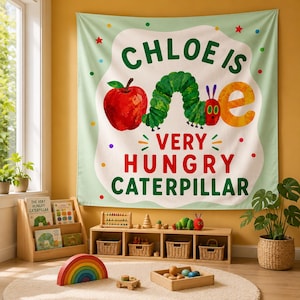 May include: A rectangular fabric wall hanging featuring a light green background with a white centre. It displays a red apple, a green caterpillar, and the text CHLOE IS ONE VERY HUNGRY CATERPILLAR in colourful letters, accented by small dots and stars.