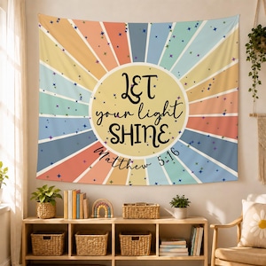Puede incluir: Un tapiz de pared rectangular con un círculo amarillo central con el texto negro Let your light SHINE Matthew 5:16. El fondo consiste en rayas radiantes en forma de sol en tonos apagados de naranja, azul, verde azulado y amarillo con estrellas.