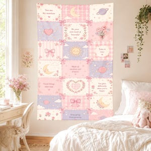 Op de afbeelding: Een rechthoekig wandkleed met een raster van pastelroze en lavendelkleurige panelen, versierd met strikken, sterren, manen, planeten en bloemen. De panelen bevatten inspirerende teksten zoals You are my sunshine, Be your own kind of beautiful, Take it easy, Made of stardust and dreams, Good things take time, You glow differently when you're happy en Keep going Keep growing.