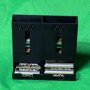 May include: A black 3D-printed battery organizer for AAA and AA batteries. The organizer has labeled slots for each battery type and a storage compartment. The batteries are visible through the slots. The background is a solid green.