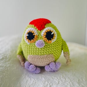 May include: A handmade, crocheted owl plushie in shades of green, red, beige, and purple. The owl has large, expressive eyes with black pupils, orange and white accents, and a red head. The plushie is sitting on a white, patterned surface.