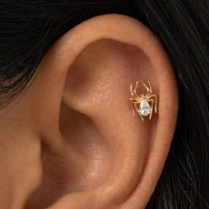 May include: A gold spider earring with a clear teardrop-shaped stone in the center worn in the upper ear cartilage and a silver spider earring with a clear teardrop-shaped stone in the center worn in the earlobe, both featuring eight legs.