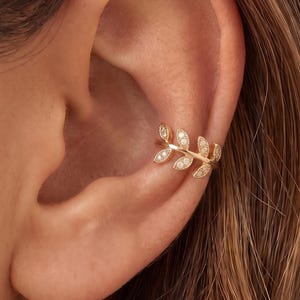 May include: A gold ear cuff featuring a delicate leaf-inspired design. The metal band is shaped like a branch with multiple small, marquise-shaped leaves, each embedded with tiny, clear, sparkling stones that catch the light along the outer edge.