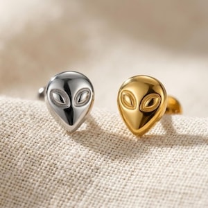 May include: Two stud earrings shaped like alien heads, one in a polished silver tone and the other in a polished gold tone. Each earring features a smooth, teardrop-shaped face with two recessed, almond-shaped eyes and a pointed chin design.