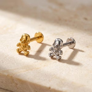 May include: Two labret stud earrings featuring a skull and crossbones design. One earring is finished in a polished gold tone, while the other is finished in a polished silver tone. Each piece includes a flat circular back and a post.
