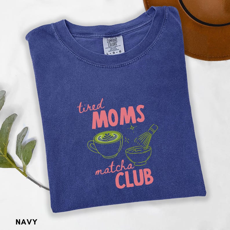 May include: Navy blue t-shirt featuring the text "tired MOMS matcha CLUB" in pink and green. The design includes a matcha latte and a whisk. The shirt is folded and has a Comfort Colors tag.