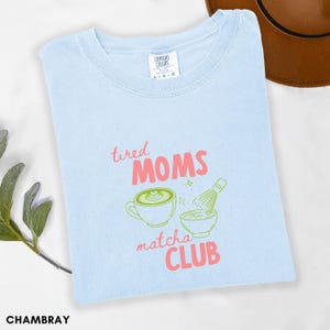 May include: Pale blue t-shirt featuring the text "tired MOMS matcha CLUB" in pink and green. The design includes a matcha latte, a bowl, and a whisk. The shirt is made of a soft material.