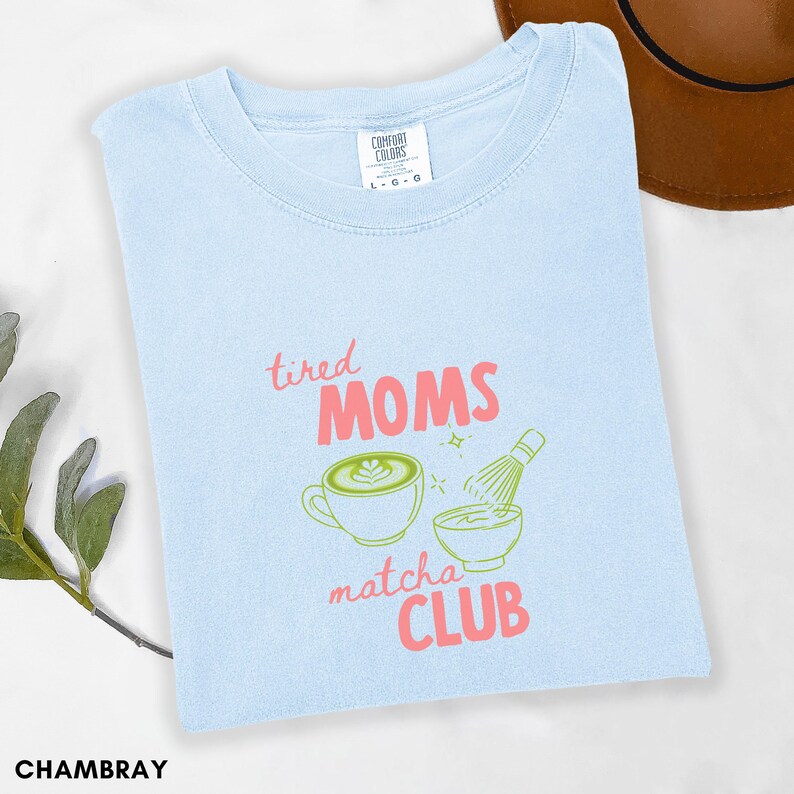 May include: Pale blue t-shirt featuring the text "tired MOMS matcha CLUB" in pink and green. The design includes a matcha latte, a bowl, and a whisk. The shirt is made of a soft material.