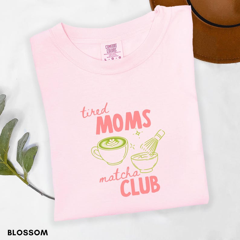 May include: A pale pink t-shirt with the text "tired MOMS matcha CLUB" in pink and green. The design includes a matcha latte and whisk graphic. The shirt is labelled "Comfort Colors" and the size is L-G.