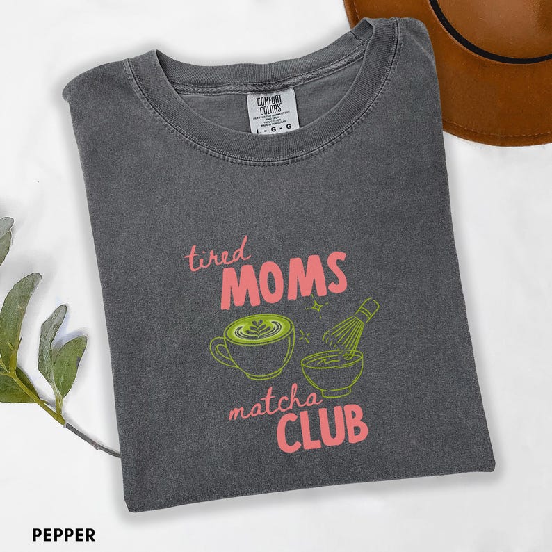 May include: A dark grey t-shirt with the words "tired MOMS matcha CLUB" in pink and green. The shirt features a coffee cup with latte art and a matcha whisk. The shirt is labelled "Pepper".